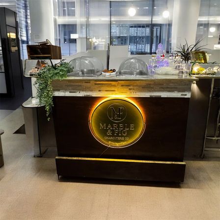 Sleek indoor charcuterie counter in an office lobby — dark wood cart with warm circular backlit logo, glass food domes, hanging greenery and illuminated displays in a modern commercial space.