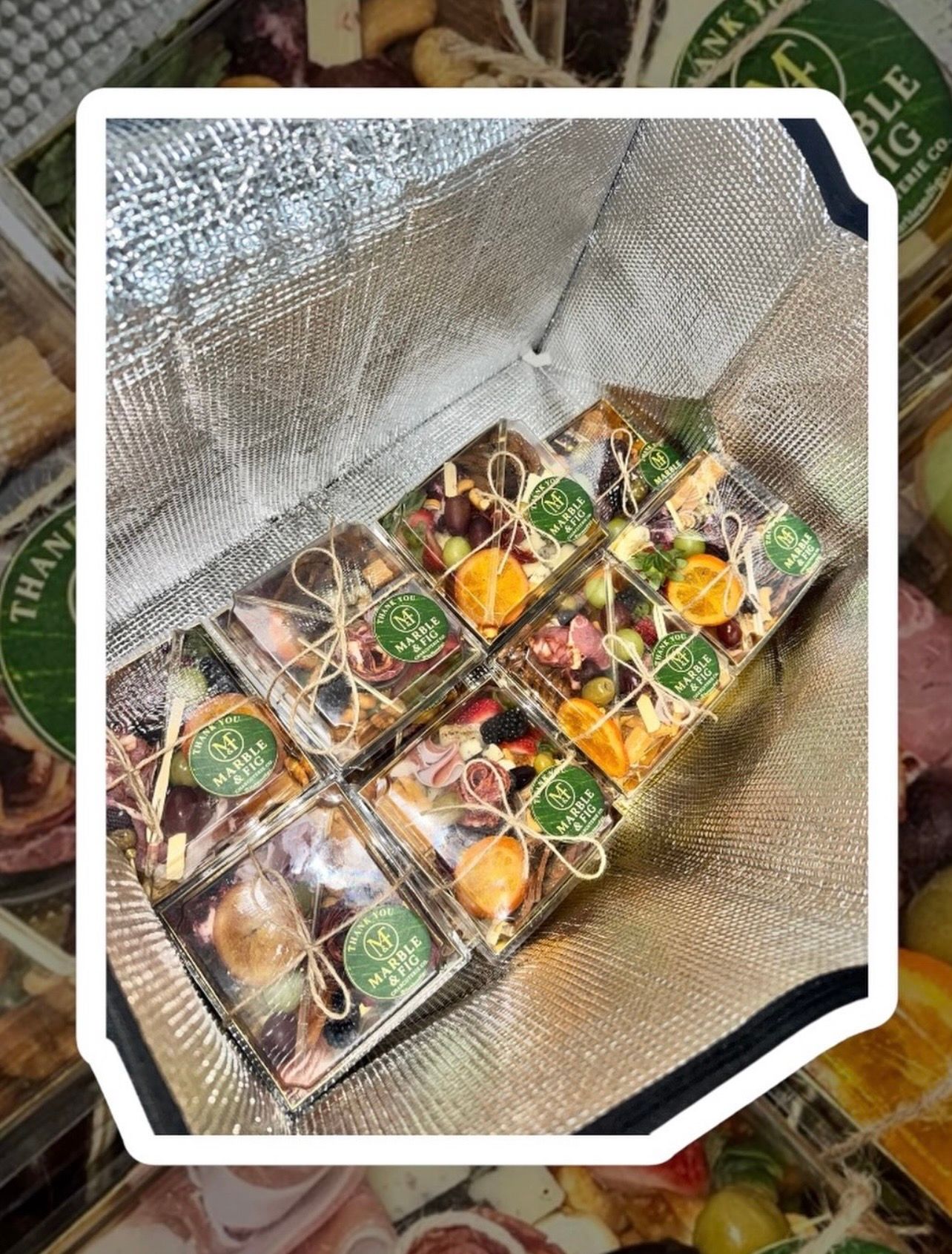 Insulated cooler bag filled with six clear mini charcuterie gift boxes — assorted fruits, cheeses, cured meats, nuts and olives tied with twine.