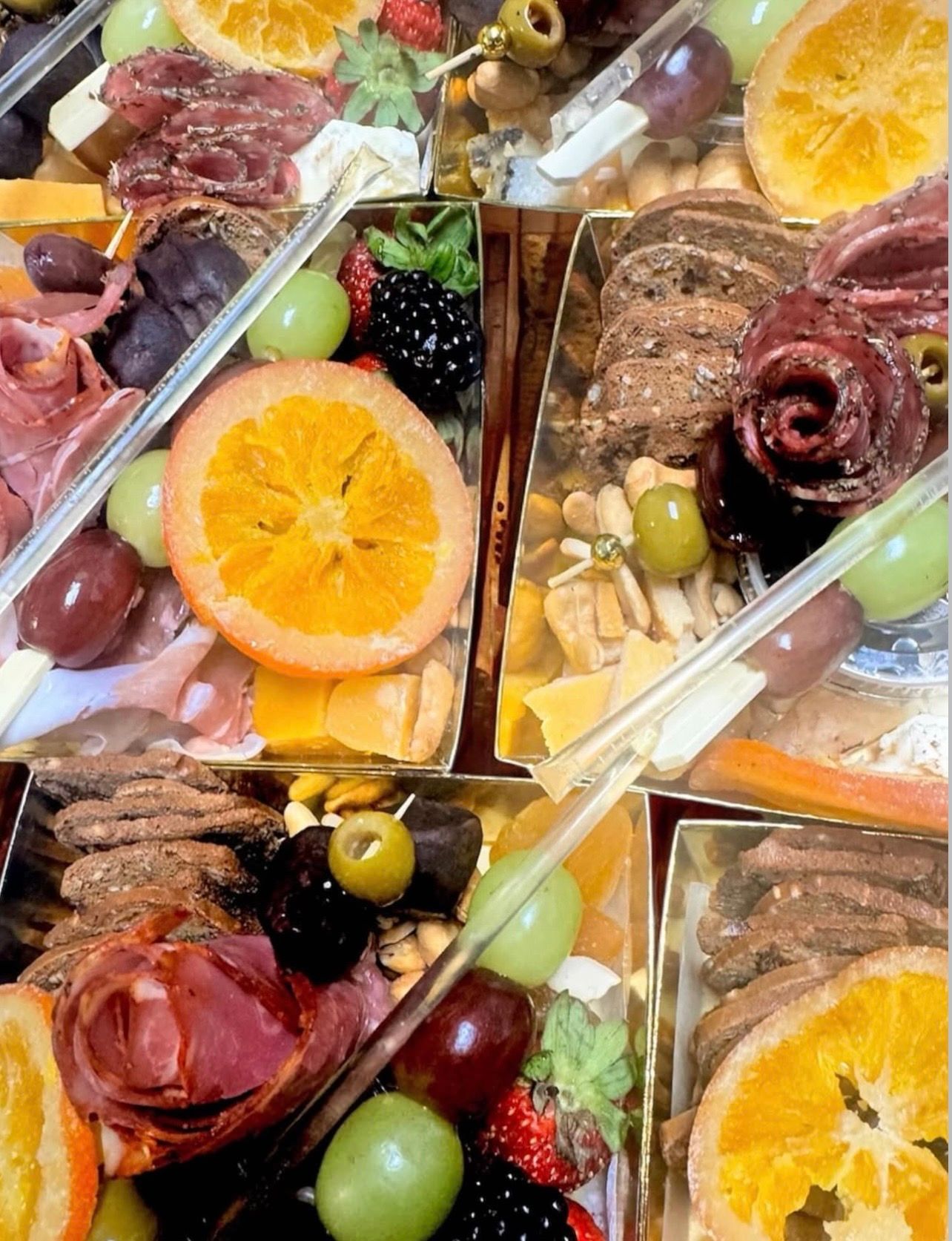 Gourmet charcuterie boxes with dried orange slices, rolled cured meats, cheeses, crackers, grapes, berries, olives, and mixed nuts