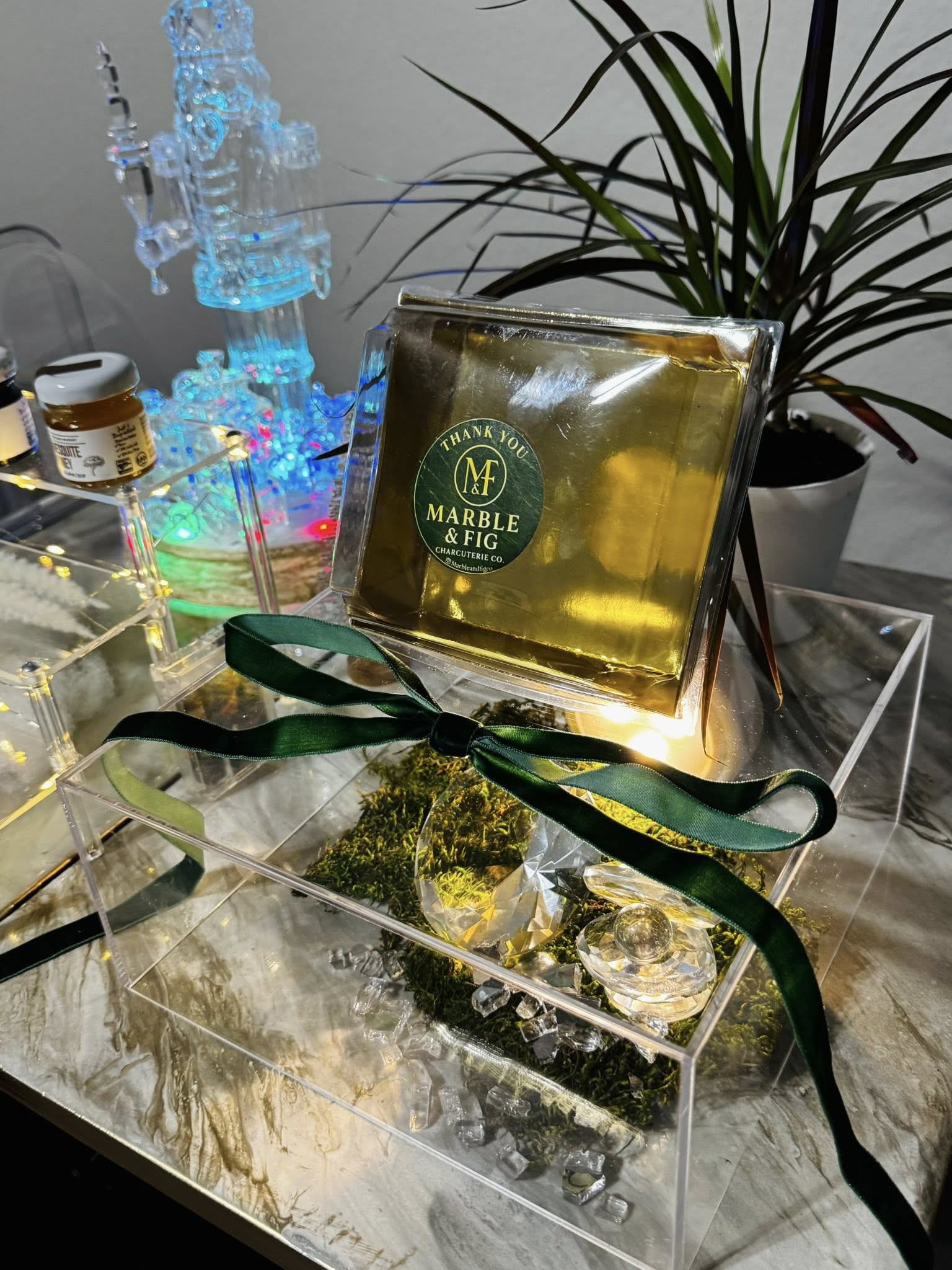 Gold-wrapped gourmet gift box with dark green ribbon on a clear acrylic display, moss and crystal accents, potted plant and colorful LED ornament on a marble-look table