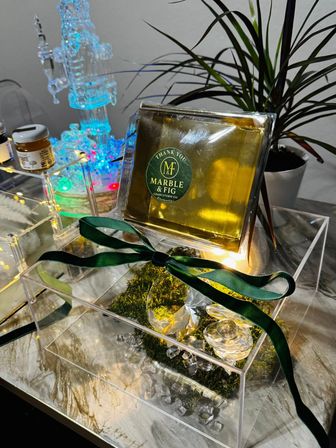 Gold-wrapped gourmet gift box with dark green ribbon on a clear acrylic display, moss and crystal accents, potted plant and colorful LED ornament on a marble-look table