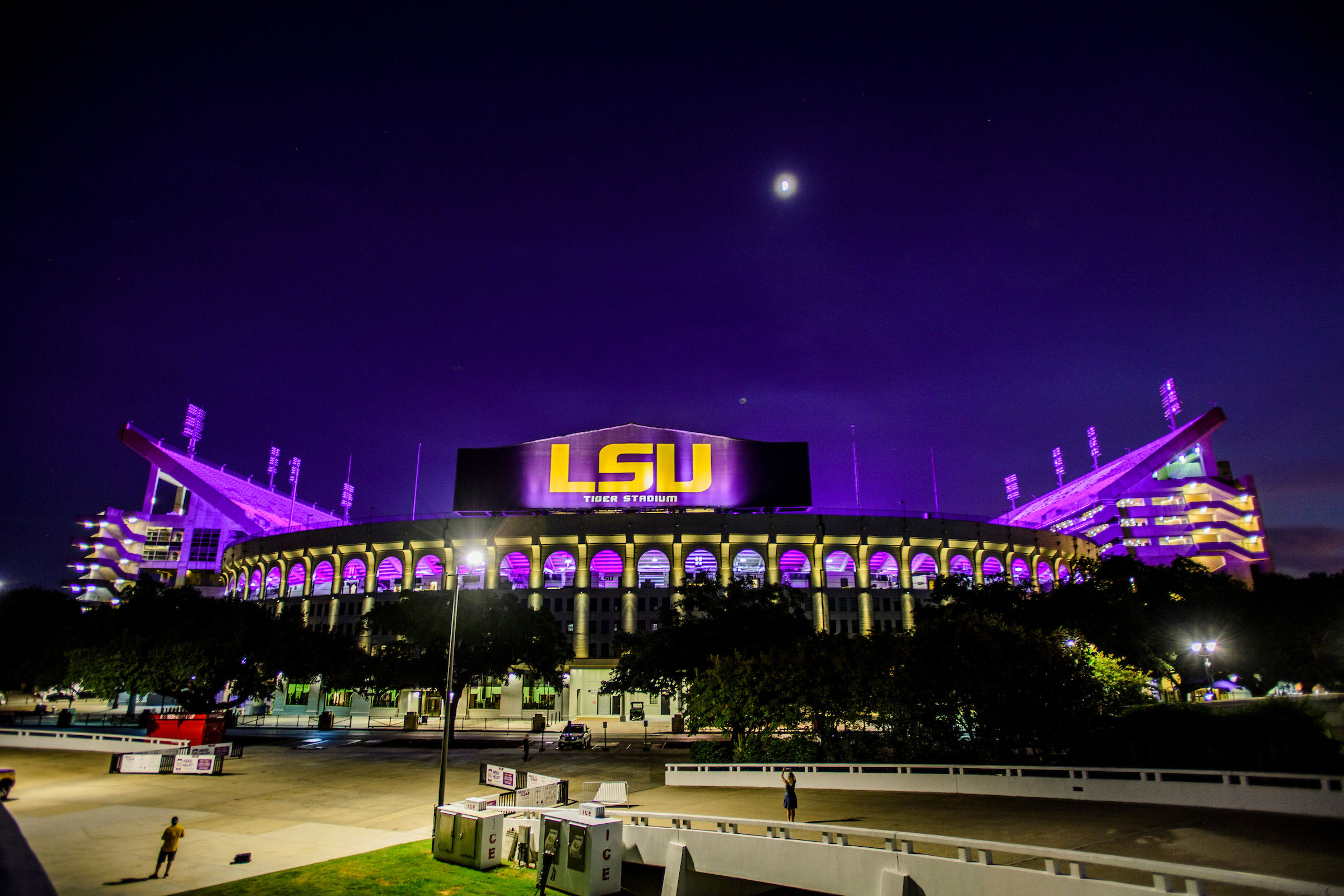 Purple-lit college football stadium glowing under a moonlit sky, arched colonnades and a giant illuminated scoreboard