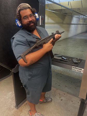 Person wearing blue hearing protection and glasses holding a folded-stock submachine-style rifle at an indoor shooting range, magazines on the bench and spent brass scattered on the concrete floor.