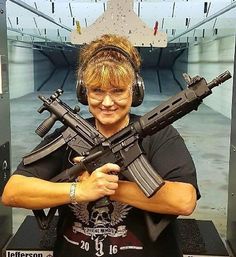 Smiling woman wearing ear protection and safety glasses at an indoor shooting range, holding two AR-style rifles crossed in front of her with a paper silhouette target downrange.
