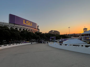 Sunset over a college football stadium with a giant purple-and-gold scoreboard, arched concourse peeking above trees, and tents and fans set up in a tailgate area.