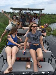Cheerful group of friends enjoying an airboat ride through green marshland, wearing sunglasses and ear protection, holding cold drinks and smiling in casual summer outfits.