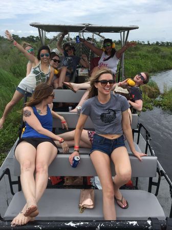 Cheerful group of friends enjoying an airboat ride through green marshland, wearing sunglasses and ear protection, holding cold drinks and smiling in casual summer outfits.