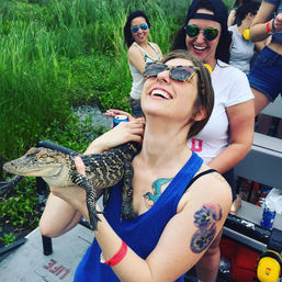 Beaming women on a swamp-tour boat holding a young alligator, wearing sunglasses and casual summer clothes amid marsh grasses