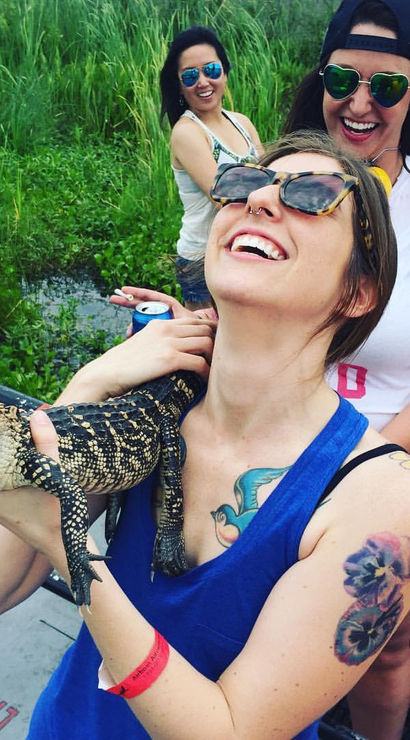 Beaming women on a swamp-tour boat holding a young alligator, wearing sunglasses and casual summer clothes amid marsh grasses