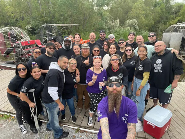 Large group of about 25 people smiling on a wooden dock next to airboats and marsh vegetation, selfie-style shot with a bearded person in purple in the foreground, casual team-outing attire and coolers