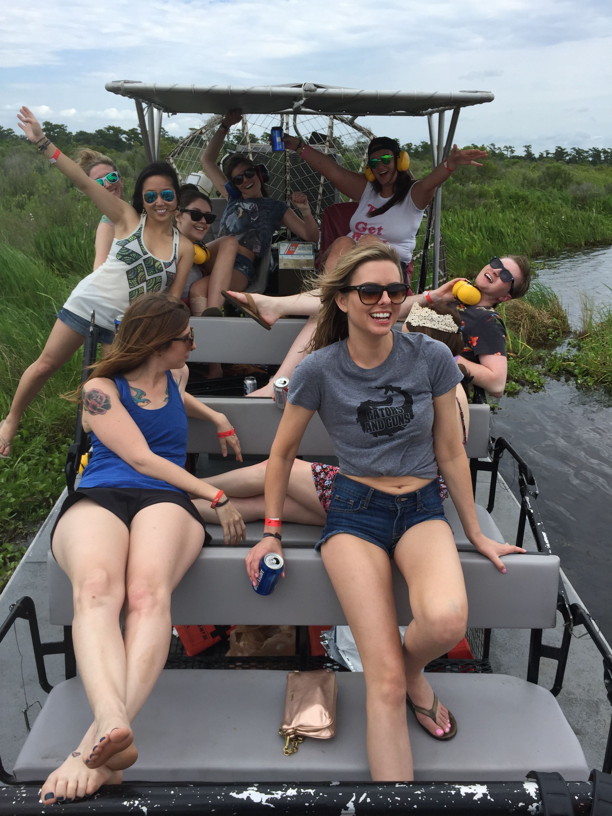 Group of young women laughing on an airboat ride through grassy Everglades marshland, wearing sunglasses and holding drinks.