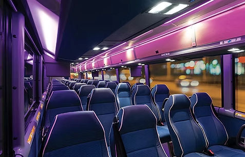 Modern coach bus interior at night with purple LED lighting, blue reclining seats, aisle view, overhead luggage racks and blurred city lights outside