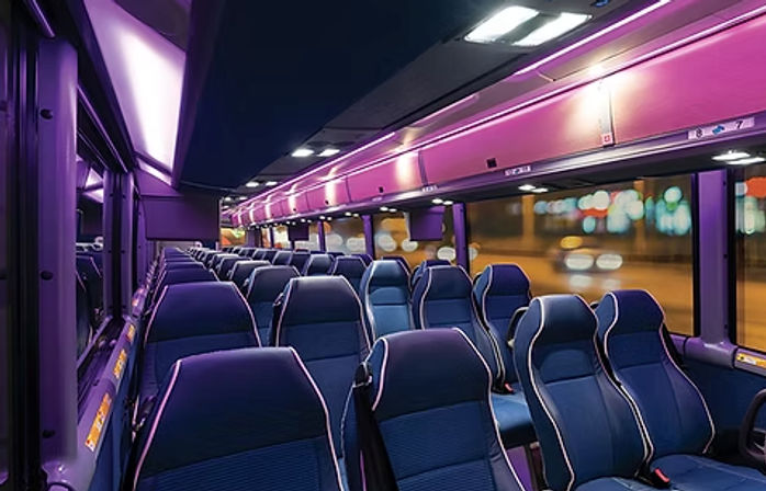 Modern coach bus interior at night with purple LED lighting, blue reclining seats, aisle view, overhead luggage racks and blurred city lights outside
