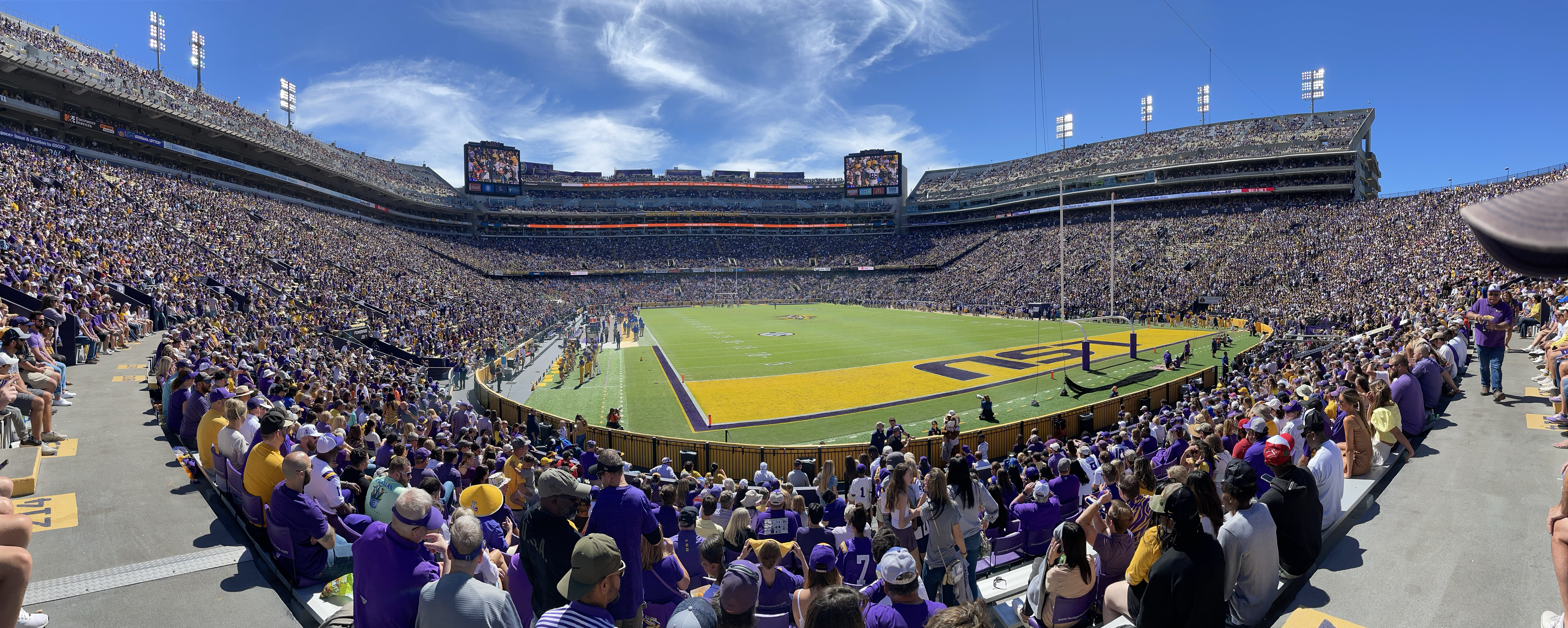 Panoramic view of a packed college football stadium on a sunny day, purple-and-gold fans filling multi-tiered stands, painted end zone and players on the field.