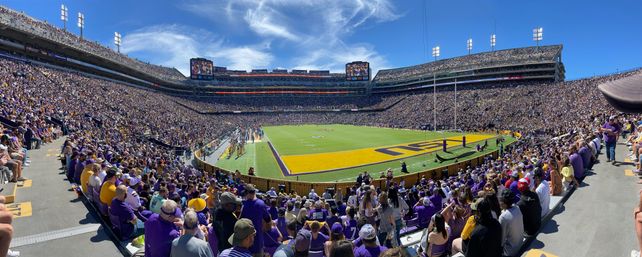 Panoramic view of a packed college football stadium on a sunny day, purple-and-gold fans filling multi-tiered stands, painted end zone and players on the field.