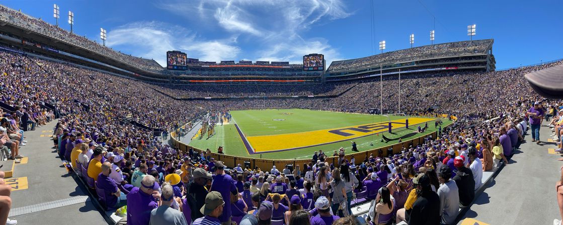 Panoramic view of a packed college football stadium on a sunny day, purple-and-gold fans filling multi-tiered stands, painted end zone and players on the field.