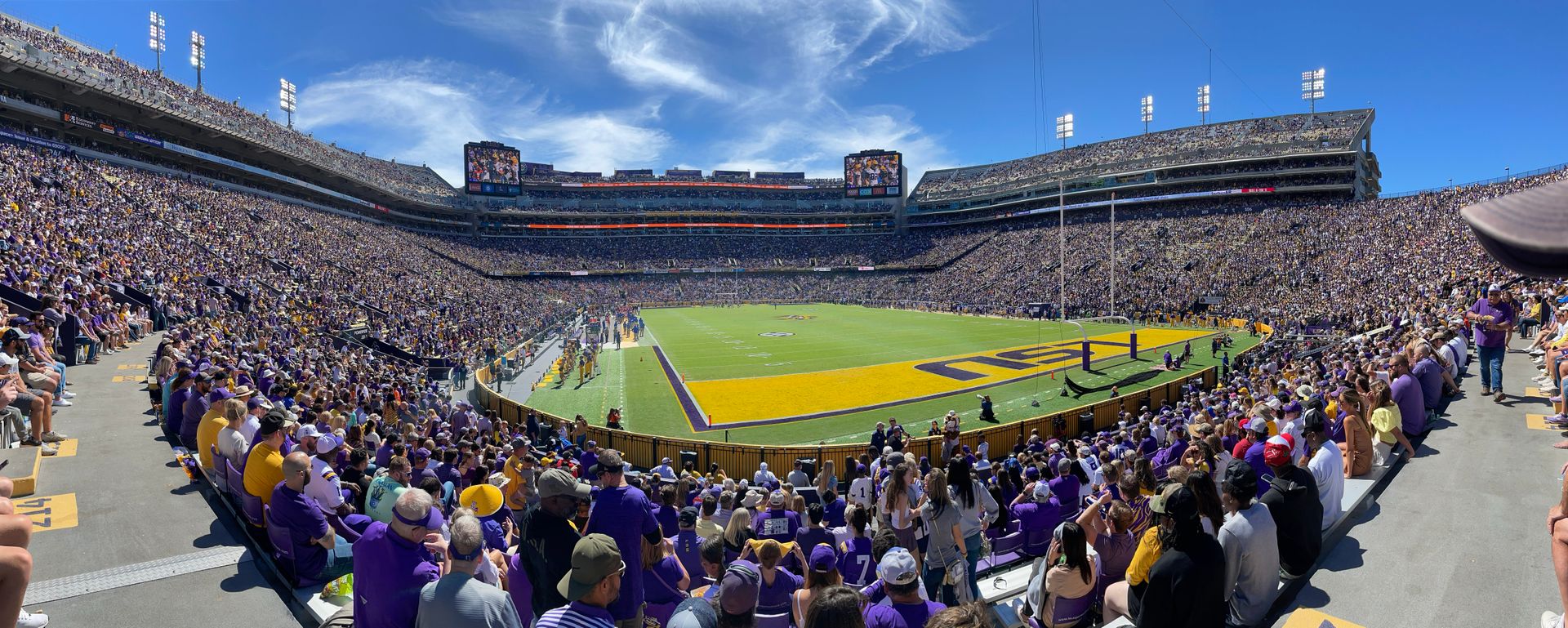 Panoramic view of a packed college football stadium on a sunny day, purple-and-gold fans filling multi-tiered stands, painted end zone and players on the field.