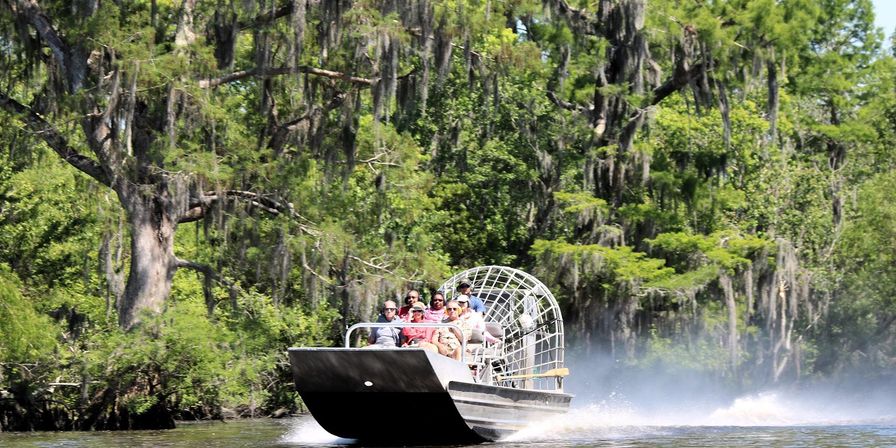 Airboat full of passengers zipping through a sunlit Louisiana cypress swamp draped in Spanish moss, leaving a spray of water behind.
