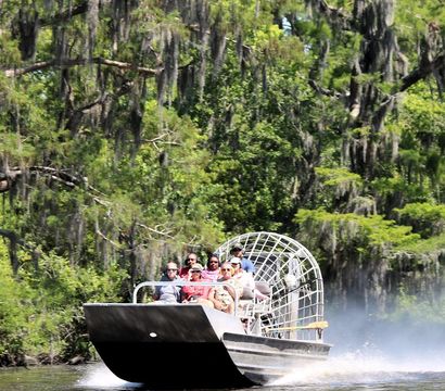Airboat full of passengers zipping through a sunlit Louisiana cypress swamp draped in Spanish moss, leaving a spray of water behind.