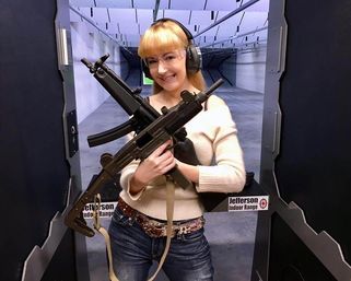 Smiling woman wearing ear protection and safety glasses at an indoor shooting range, holding two submachine-style firearms across her chest while dressed in a sweater and jeans.