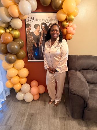 Smiling woman in blush satin pajamas poses in a cozy living room beside a peach-and-gold balloon arch and a 'Waiting to Exhale' poster next to a gray sofa