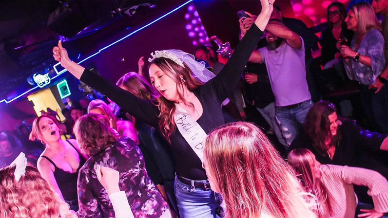 Veiled bride-to-be wearing a sash dancing exuberantly with friends in a crowded neon-lit nightclub bachelorette party.