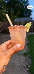 Hand holding an icy pink cocktail in a plastic cup with a chili-salt rim, lime wedge and paper straw, blurred backyard fence and trees in the background.