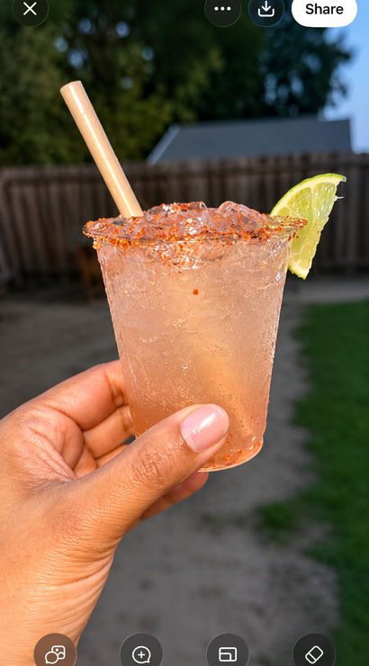 Hand holding an icy pink cocktail in a plastic cup with a chili-salt rim, lime wedge and paper straw, blurred backyard fence and trees in the background.