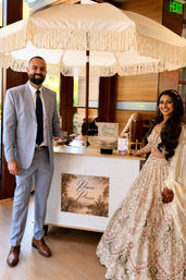 Smiling bride in an embroidered lehenga and groom in a light-gray suit stand by a fringed-umbrella dessert cart at an indoor wedding reception.