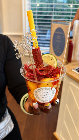 Colorful Mexican-style fruit cup: mango chunks with chamoy, chili powder and shredded tamarind candy, lime slice and tamarind straw in a clear plastic cup held by hand.