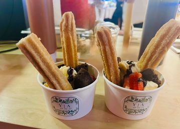 Two dessert cups on a wooden table with sugar-coated churros, vanilla ice cream, brownie bites, sliced strawberries and chocolate drizzle — a colorful cafe-style treat.