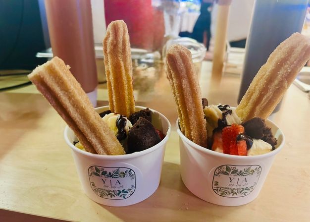 Two dessert cups on a wooden table with sugar-coated churros, vanilla ice cream, brownie bites, sliced strawberries and chocolate drizzle — a colorful cafe-style treat.
