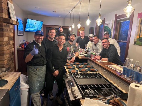 Group of friends in a cozy home kitchen gathered around a stove and island with multiple plated pancakes; a chef wearing a glove gives a thumbs-up under hanging pendant lights while others smile, with bottles and a TV showing sports in the background.