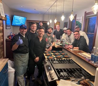 Group of friends in a cozy home kitchen gathered around a stove and island with multiple plated pancakes; a chef wearing a glove gives a thumbs-up under hanging pendant lights while others smile, with bottles and a TV showing sports in the background.