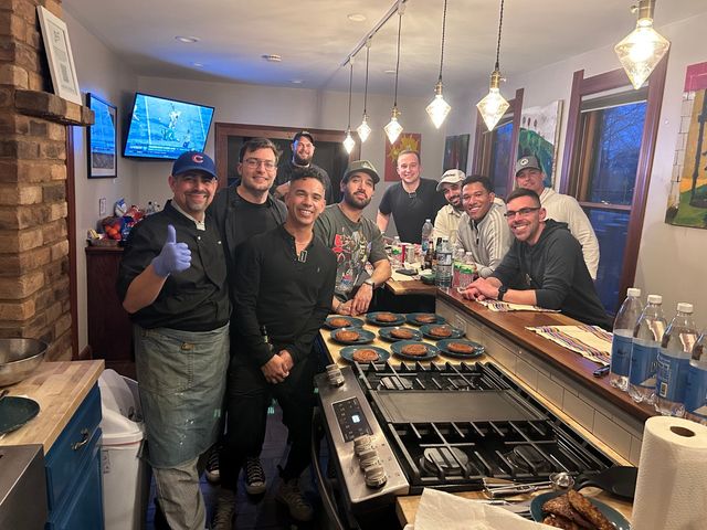 Group of friends in a cozy home kitchen gathered around a stove and island with multiple plated pancakes; a chef wearing a glove gives a thumbs-up under hanging pendant lights while others smile, with bottles and a TV showing sports in the background.