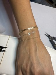 Close-up of a wrist wearing three delicate stacked bracelets — a gold disc-link chain, a silver chain with a small round charm, and a thin gold chain — resting on a white jewelry work surface with pliers nearby.