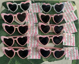 Array of heart-shaped pink and white sunglasses embellished with pearls, rhinestones and letter beads, arranged with pink-striped cards reading 'Girls just want to have sun in Saint Lucia' — playful tropical bachelorette party favors.