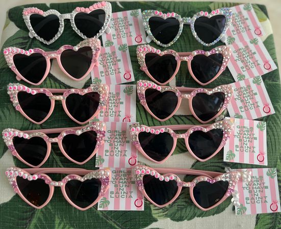 Array of heart-shaped pink and white sunglasses embellished with pearls, rhinestones and letter beads, arranged with pink-striped cards reading 'Girls just want to have sun in Saint Lucia' — playful tropical bachelorette party favors.