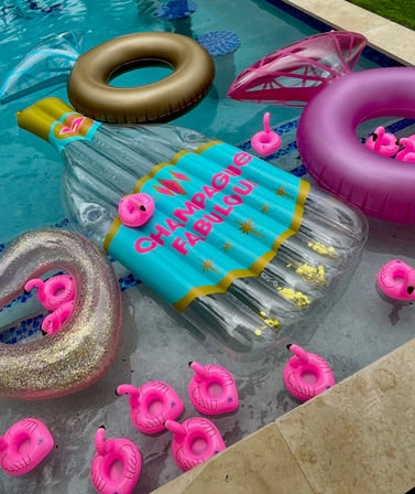 Backyard pool with a large inflatable champagne bottle float labeled "CHAMPAGNE FABULOUS", pink flamingo drink-ring holders, gold-glitter and purple pool rings in clear blue water — playful summer party vibe.
