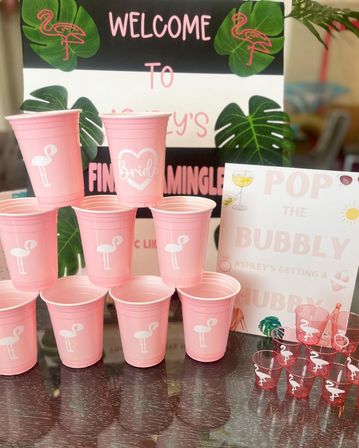 Pink tropical-themed bachelorette party setup with a pyramid of pink plastic cups featuring white flamingo and a 'Bride' cup, matching flamingo shot cups, and 'Welcome' and 'Pop the Bubbly' signage with monstera leaf accents.
