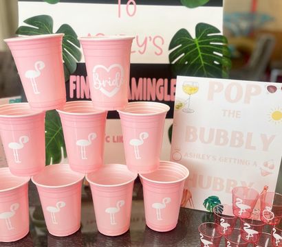 Pink tropical-themed bachelorette party setup with a pyramid of pink plastic cups featuring white flamingo and a 'Bride' cup, matching flamingo shot cups, and 'Welcome' and 'Pop the Bubbly' signage with monstera leaf accents.
