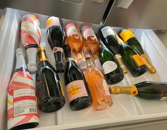 Refrigerator drawer filled with assorted sparkling wine and champagne bottles—rosé and brut varieties with colorful labels and foil caps, chilled and laid on their sides, ready for a celebration.