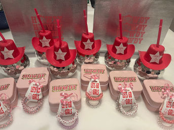 Pink western-themed party favors on a table: red mini cowboy-hat tumblers with straws perched on mirrored disco-ball bases, pink boxes stamped 'Howdy', and stacks of pink beaded bracelets with 'Let's Go Girls' tags.