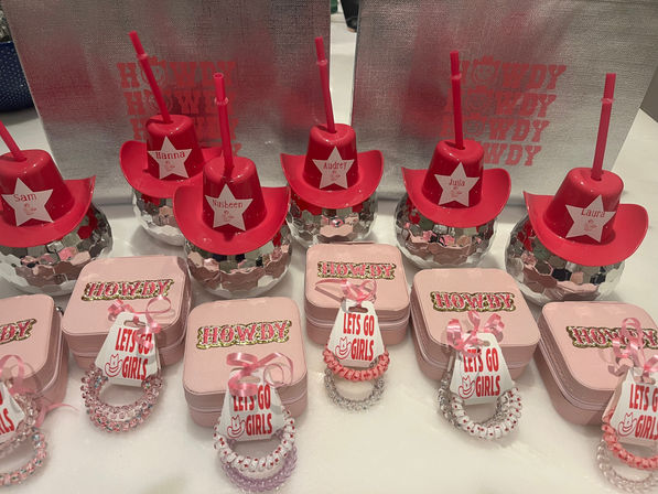 Pink western-themed party favors on a table: red mini cowboy-hat tumblers with straws perched on mirrored disco-ball bases, pink boxes stamped 'Howdy', and stacks of pink beaded bracelets with 'Let's Go Girls' tags.