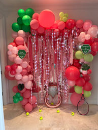 Indoor playful party photo backdrop with pink and red balloon arch, shimmering star fringe curtain, neon yellow tennis balls scattered on floor, black-and-pink tennis racket, green balloon clusters and metallic ring prop — tennis-themed balloon decor.