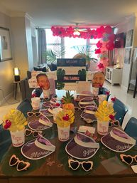 Flamingo-themed bachelorette party setup in a bright apartment living room, featuring a table with personalized visors and cups, heart-shaped sunglasses, tropical leaf placemats, pineapple centerpiece, gold leaf decorations, large face cutouts, and a pink balloon arch with a 'Final Flamingle' welcome sign.