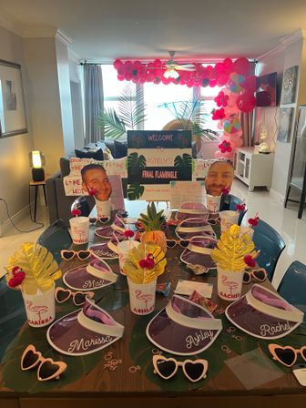 Flamingo-themed bachelorette party setup in a bright apartment living room, featuring a table with personalized visors and cups, heart-shaped sunglasses, tropical leaf placemats, pineapple centerpiece, gold leaf decorations, large face cutouts, and a pink balloon arch with a 'Final Flamingle' welcome sign.
