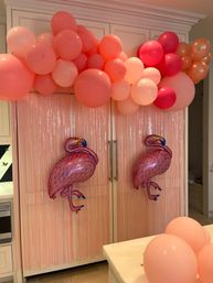 Kitchen party backdrop with coral, blush and rose-gold balloon garland over cabinet doors, pink fringe curtain, two shiny pink flamingo foil balloons and pink balloons on the countertop.