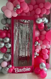 Pink doll-box photo booth display with sparkly silver reflective interior, framed by a balloon arch of hot pink, light pink and metallic silver balloons with mirrored disco-ball accents.