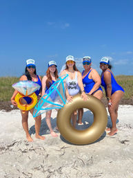 Five women in matching blue swimsuits and trucker hats posing on a sunny sandy beach for a bachelorette celebration, holding a gold inner tube and oversized diamond-ring inflatables.